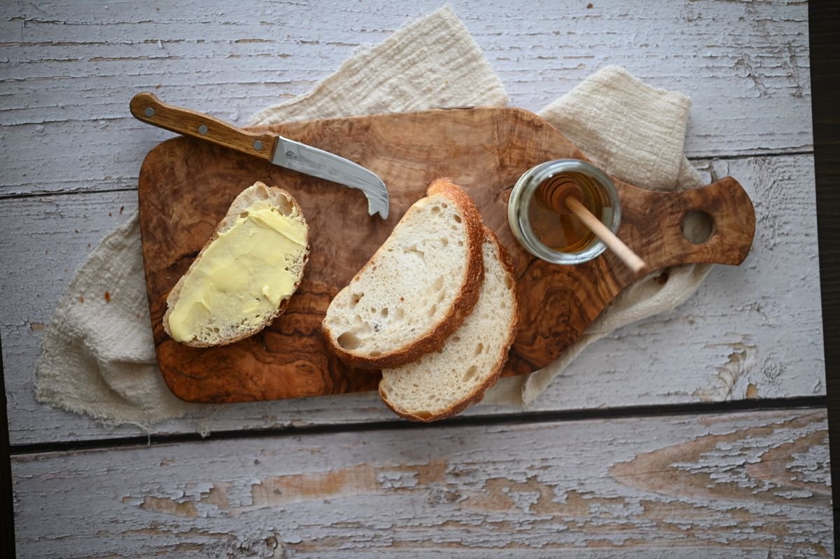 Composição rústica com fatias de pão, manteiga caseira passada com faca de cabo de madeira e pote de mel em tábua de madeira