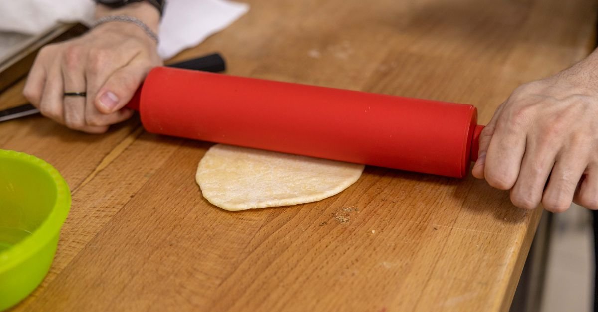 Mãos abrindo massa de pastel com rolo vermelho em bancada de madeira