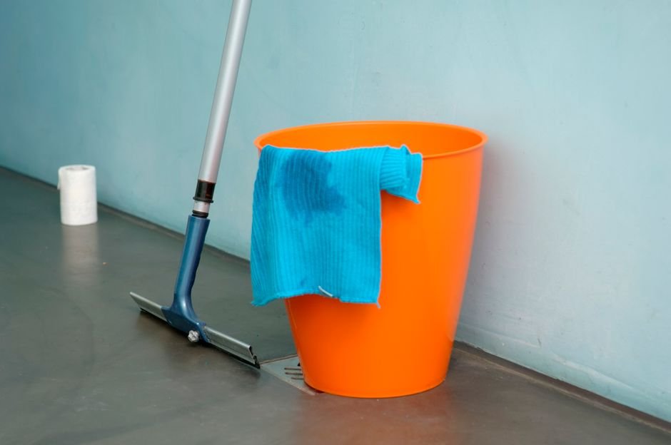 Balde laranja com pano azul e rodo encostado na parede para limpeza do piso