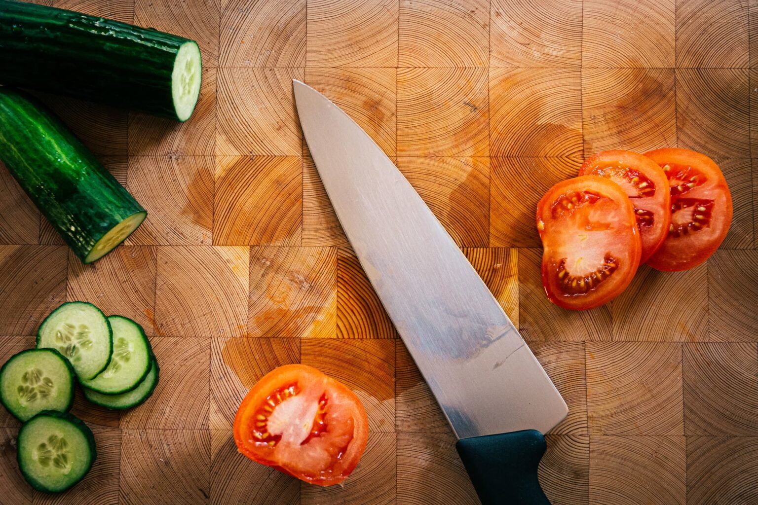 Mãos cortando pepino e tomate sobre tábua de corte de madeira na bancada da cozinha: como escolher tábua de corte