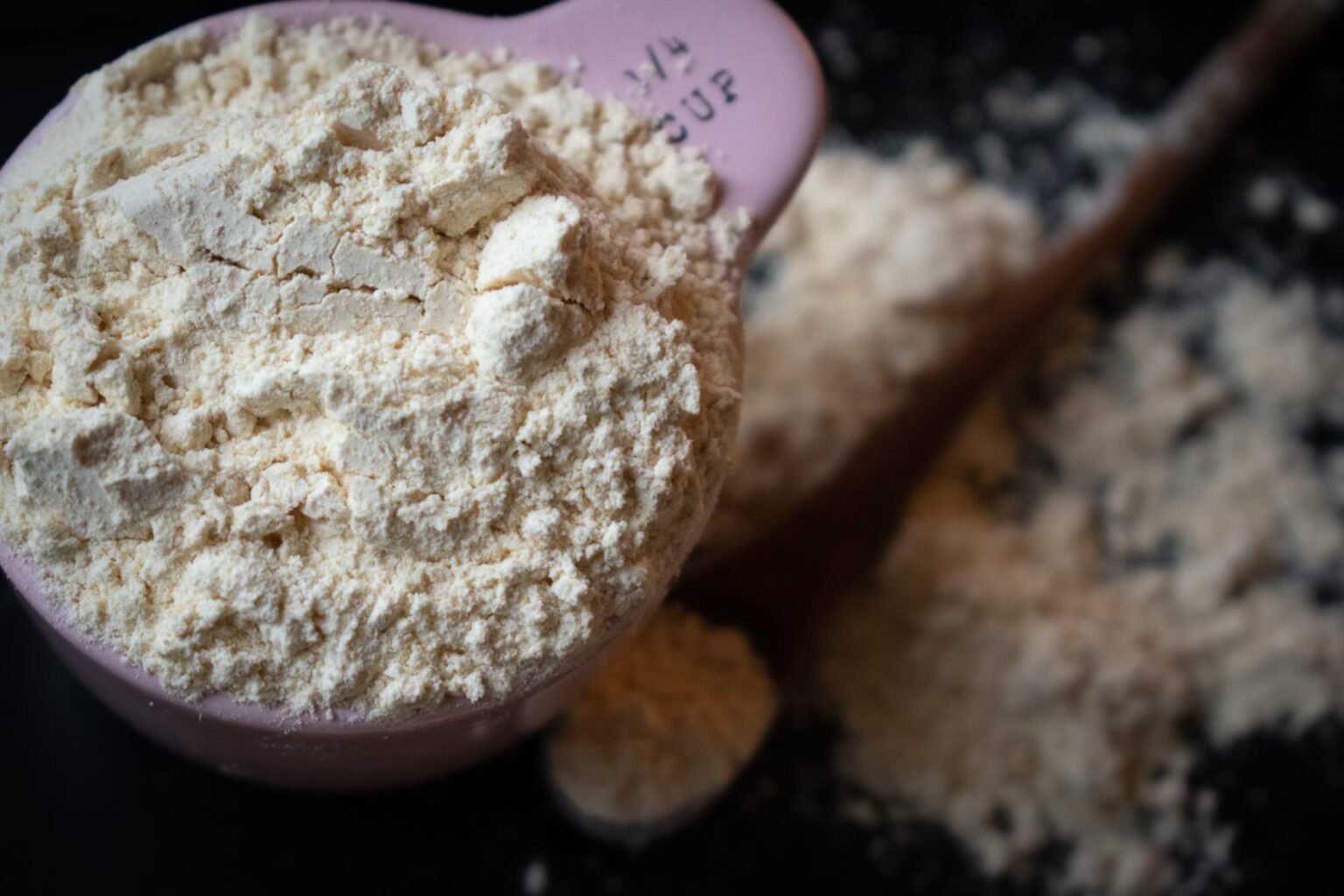 Xícara medidora transbordando de farinha de trigo sobre superfície escura: tabela de medidas na cozinha em gramas para não compactar o ingrediente